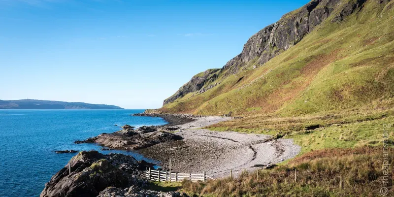 A photograph of a Scottish sea loch on a sunny day, with a fence in the foreground, a pebbly bay behind it, and rocky cliffs descending to the sea in the distance