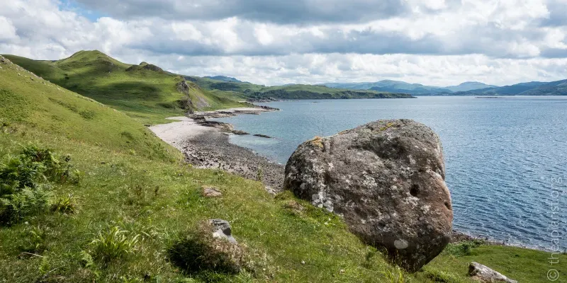 Photo of a Scottish sea loch with a pebbly beach in the distance and a large boulder in the foreground.