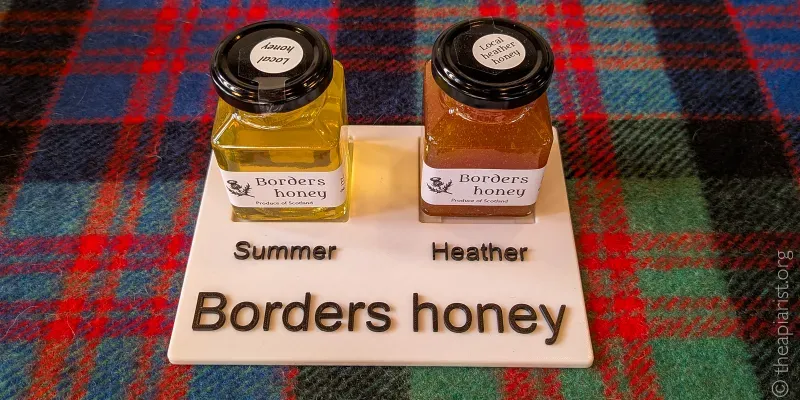 Two jars of honey on a white platform. Both jars are labelled 'Borders honey', and one is summer honey and the other is heather honey. The white platform (on a tartan cloth) indicates the type of honey.
