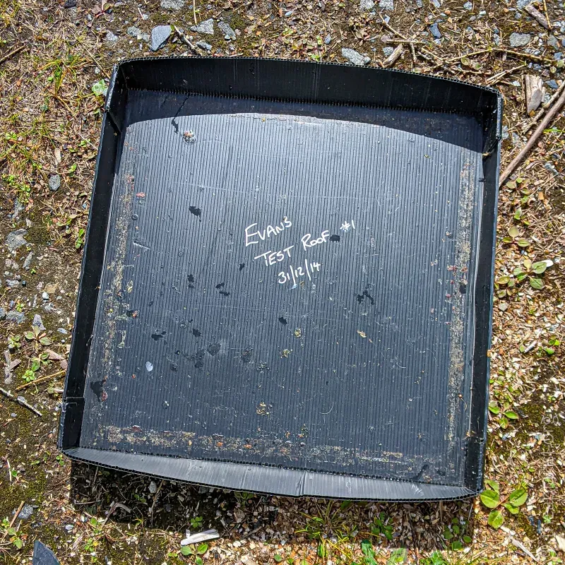A black, plastic, folded bee hive roof lying on the ground, with the works Test Roof #1 and the a date written on the underside.