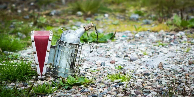 An old beekeeping smoker, with a cork in the spout, standing on gravel.