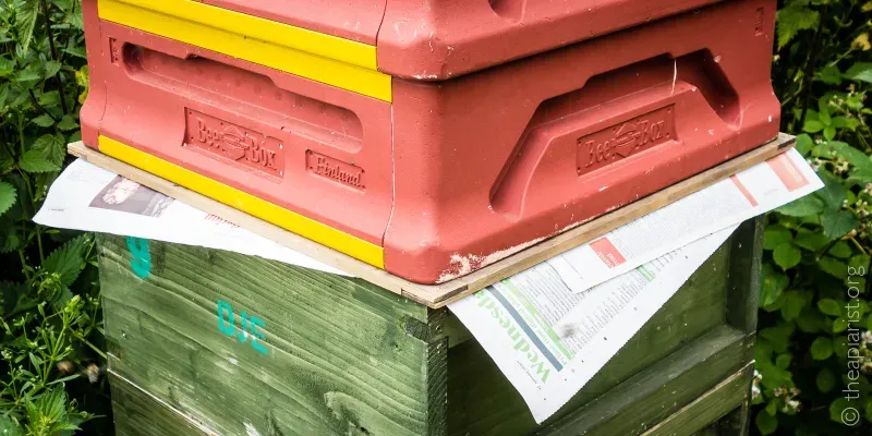 A photo of two different types of bee hive box stacked on top of each other, with newspaper in between.
