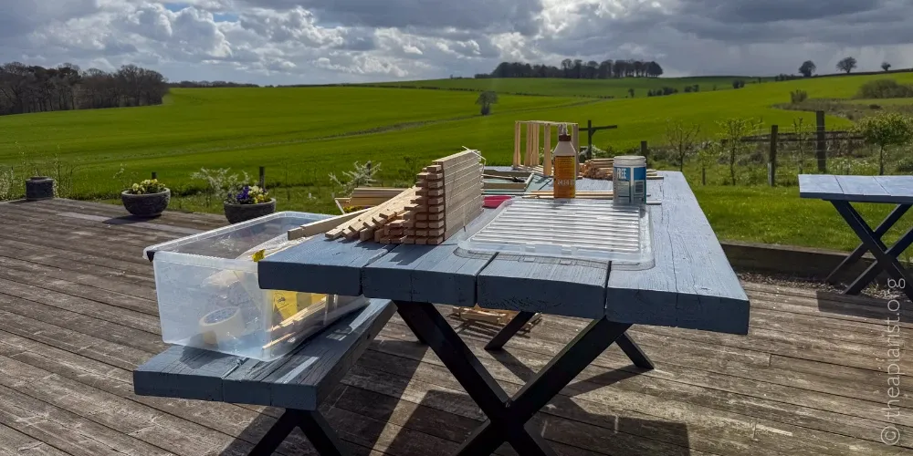 A photograph of an outdoor table covered with beekeeping frame making equipment, with a distant view of open farmland.