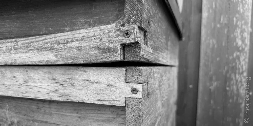 Two wooden bee hive boxes on top of each other, showing detail of the junction between them. The upper box is warped and there is a gap between them.