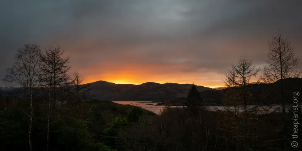 A view across a Scottish loch at dawn in winter, with the sun rising over distant hills, but the rest of the image in shadow