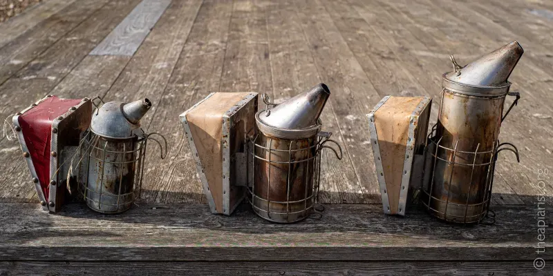 Three beekeeping smokers of different sizes in a line, on wooden decking