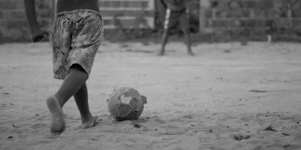 A B&W photograph of a boy playing with a tattered football on a beach in Brazil.