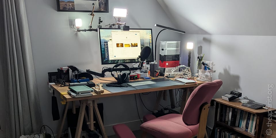 A home office Zoom setup in an attic room with a sloped white ceiling. A wooden trestle desk holds a large monitor, condenser microphone on a boom arm, and over-ear headphones.