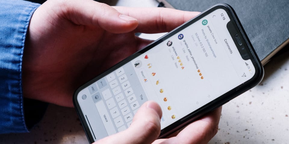 A person holds a smartphone with both hands, typing on the on-screen keyboard while viewing a social media comments section filled with emoji reactions.